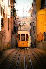 An old-fashioned yellow tram ascends a steep, narrow street lined with buildings. People are visible inside the tram, and some are standing outside, watching. The buildings on either side are adorned with graffiti, and some have laundry hanging from balconies. The warm sunlight creates a vibrant and inviting atmosphere.