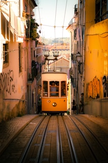 An old-fashioned yellow tram ascends a steep, narrow street lined with buildings. People are visible inside the tram, and some are standing outside, watching. The buildings on either side are adorned with graffiti, and some have laundry hanging from balconies. The warm sunlight creates a vibrant and inviting atmosphere.