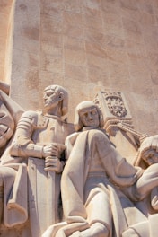 Sculptural relief depicting historical figures carved in a monumental stone facade, with detailed expressions and clothing. One figure holds a sword, while another has an emblem with a cross.