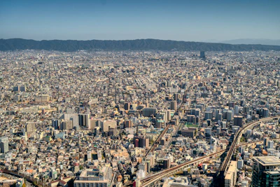 An expansive urban landscape stretches towards the horizon, showcasing a densely packed city with numerous buildings ranging from low-rises to high-rise structures. The skyline is surrounded by distant mountains under a clear blue sky. Multiple roads and rail lines weave through the cityscape, indicating a well-connected infrastructure.