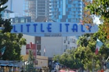 An archway sign with bold blue letters reads 'Little Italy San Diego', surrounded by lush green trees and urban buildings. The area appears vibrant with an outdoor dining scene, featuring umbrellas and flags.