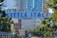 An archway sign with bold blue letters reads 'Little Italy San Diego', surrounded by lush green trees and urban buildings. The area appears vibrant with an outdoor dining scene, featuring umbrellas and flags.