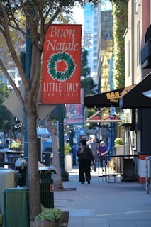 A street scene in an urban area with a prominent red sign reading 'Buon Natale' and 'Little Italy San Diego' with a green wreath image. Sidewalks are lined with trees and shops, including a bar and a pizzeria. People are walking along the street under clear blue skies, giving a festive, holiday atmosphere.