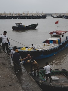 Workers are on a dock interacting with a boat heavily loaded with goods, likely fish or seafood, as it is moored beside the dock. A small boat with a flag is floating nearby in the harbor, which is filled with numerous similar ships in the background under a cloudy sky.