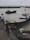 Workers are on a dock interacting with a boat heavily loaded with goods, likely fish or seafood, as it is moored beside the dock. A small boat with a flag is floating nearby in the harbor, which is filled with numerous similar ships in the background under a cloudy sky.