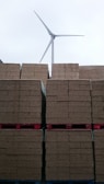 Stacks of brown bricks are neatly arranged on red pallets, with a wind turbine standing tall in the background against a cloudy sky.