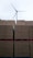 Stacks of brown bricks are neatly arranged on red pallets, with a wind turbine standing tall in the background against a cloudy sky.