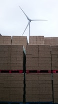 Stacks of brown bricks are neatly arranged on red pallets, with a wind turbine standing tall in the background against a cloudy sky.