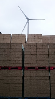Stacks of brown bricks are neatly arranged on red pallets, with a wind turbine standing tall in the background against a cloudy sky.