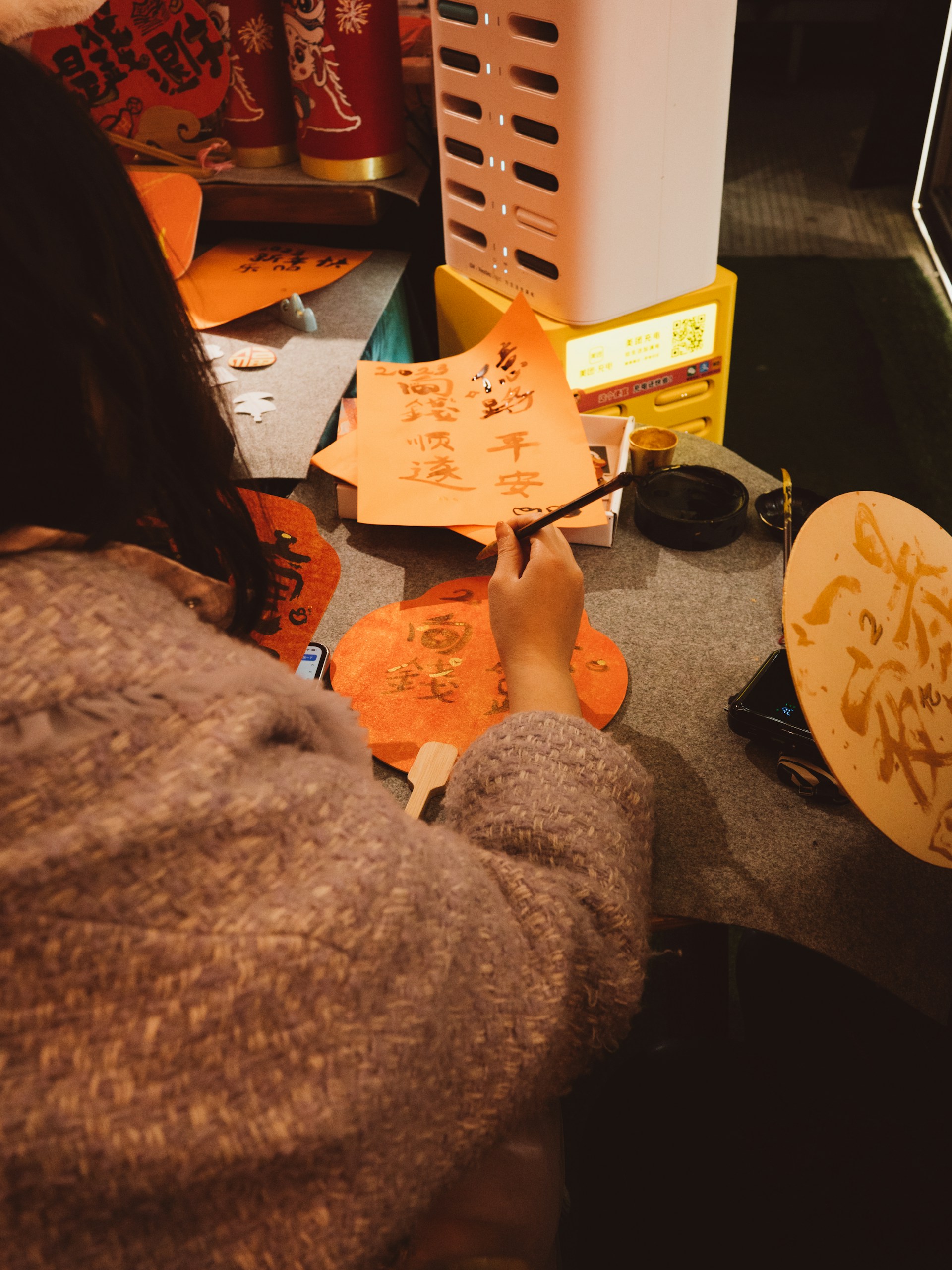 A person is engaged in calligraphy, writing Chinese characters on orange and red paper with a brush. The setting appears to be indoors with a modern heater in the background. Traditional decorations and tools are scattered on the table, indicating a festive or cultural activity.