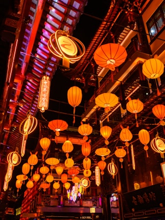 A vibrant street scene filled with numerous glowing orange and yellow lanterns hanging above. Traditional architecture and neon signs are visible, creating a lively and culturally rich atmosphere.