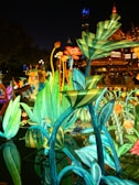 Illuminated plants and flowers created from colorful lights and structures during a night festival. The vibrant installation reflects in a body of water, with a traditional architectural building and glowing street lamps visible in the background.