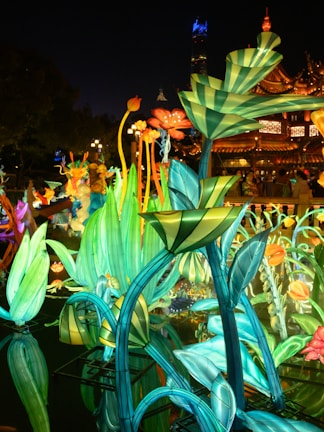 Illuminated plants and flowers created from colorful lights and structures during a night festival. The vibrant installation reflects in a body of water, with a traditional architectural building and glowing street lamps visible in the background.