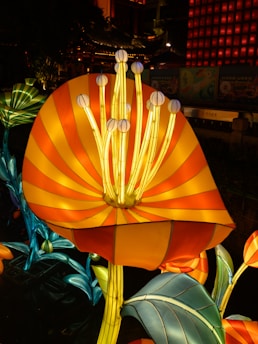 Brightly lit, large-scale floral sculptures with vibrant colors, including orange, yellow, and blue, glow in a nighttime setting. The flowers exhibit a lantern-like design with intricate details in their petals and stems.