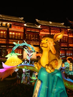 A vibrant and colorful display featuring a large, whimsical illuminated figure resembling a fantasy creature with exaggerated features. The creature sits atop a brightly lit base surrounded by luminous flowers, creating a festive and magical atmosphere. In the background, a traditional style building is adorned with warm lights, which enhances the overall enchanting ambiance.