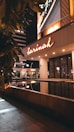 A nighttime scene at the entrance of a modern building with a prominent illuminated sign reading 'Sarinah'. The building features a reflective water pool in front and is surrounded by lush green foliage. The area is well-lit, creating contrasts of light and shadow.