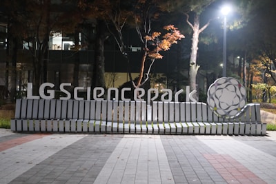A modern bench with a sleek design featuring the text 'LG Sciencepark' is situated in an outdoor setting. The bench is illuminated by a streetlight and is surrounded by trees with autumn foliage. A large, decorative structure resembling a molecule sits to the right of the bench. The ground is paved with interlocking tiles in various shades of gray and red.
