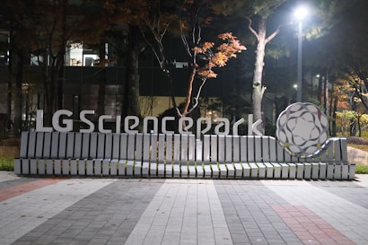 A modern bench with a sleek design featuring the text 'LG Sciencepark' is situated in an outdoor setting. The bench is illuminated by a streetlight and is surrounded by trees with autumn foliage. A large, decorative structure resembling a molecule sits to the right of the bench. The ground is paved with interlocking tiles in various shades of gray and red.