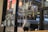 A view from the inside of a café showing a glass door with signage including 'LA BAB' and symbols for WiFi and CCTV. Reflections of interior lighting are visible on the glass. Outside, blurred outlines of people and city lights create a bustling nighttime city street scene. Umbrellas are placed on the left side, suggesting a rainy setting.