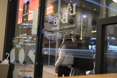 A view from the inside of a caf&eacute; showing a glass door with signage including 'LA BAB' and symbols for WiFi and CCTV. Reflections of interior lighting are visible on the glass. Outside, blurred outlines of people and city lights create a bustling nighttime city street scene. Umbrellas are placed on the left side, suggesting a rainy setting.