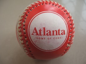 A red and white baseball features the word 'Atlanta' prominently in red text, along with the phrase 'Home of Coke' underneath. The ball is neatly stitched and its colors are vivid against a neutral background.