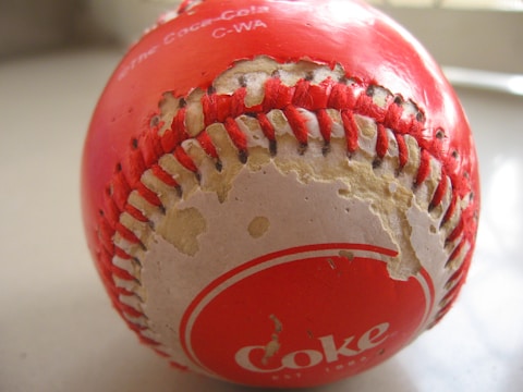 A worn out baseball with visible red stitching and a partially peeled coating. The surface shows signs of wear and tear with exposed inner material. The brand 'Coke' is printed in white on the red sections of the ball.