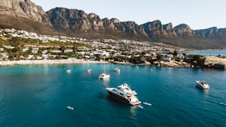 An aerial view of a coastal area with clear blue waters and several yachts and boats scattered across the bay. Luxurious houses are nestled along the shoreline, leading up to a range of rugged, majestic mountains in the background. The beach and waterfront areas suggest a lively atmosphere with people likely enjoying leisure activities.