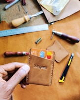 A hand holding a brown leather cardholder containing two colorful cards, with the brand name embossed on the front. The background features various leatherworking tools such as a ruler, crafting knife, and awls on a brown leather surface.