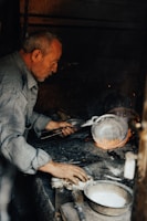 An older man works intently over a hot forge, holding metalwork tools and focusing on a piece of metal that glows with heat. The surroundings are dimly lit, with a rustic atmosphere. A container with a white substance and a brush are present on the workbench.