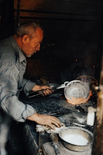 An older man works intently over a hot forge, holding metalwork tools and focusing on a piece of metal that glows with heat. The surroundings are dimly lit, with a rustic atmosphere. A container with a white substance and a brush are present on the workbench.