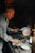 An older man works intently over a hot forge, holding metalwork tools and focusing on a piece of metal that glows with heat. The surroundings are dimly lit, with a rustic atmosphere. A container with a white substance and a brush are present on the workbench.