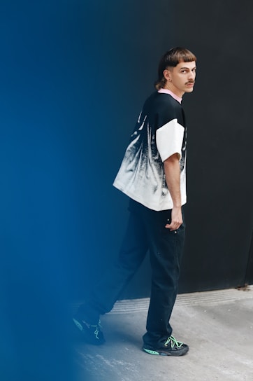 A person with short hair and a mustache is wearing a fashionable oversized shirt with a graphic design and dark pants. The setting is minimalistic with a blue and black background, adding a modern artistic touch.