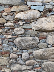 A textured stone wall composed of variously sized and shaped stones, ranging from large rounded boulders to smaller flat pieces. The stones are mostly in natural shades of gray, brown, and tan, with some hints of red and blue.