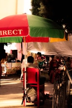 A lively street market scene with a large red and green umbrella providing shade to several people. A child dressed in colorful clothing plays nearby, while adults sit on chairs under the canopy. The surroundings are adorned with patterned textiles and various market stalls.