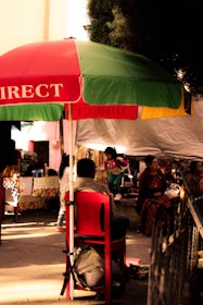 A lively street market scene with a large red and green umbrella providing shade to several people. A child dressed in colorful clothing plays nearby, while adults sit on chairs under the canopy. The surroundings are adorned with patterned textiles and various market stalls.