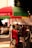 A lively street market scene with a large red and green umbrella providing shade to several people. A child dressed in colorful clothing plays nearby, while adults sit on chairs under the canopy. The surroundings are adorned with patterned textiles and various market stalls.