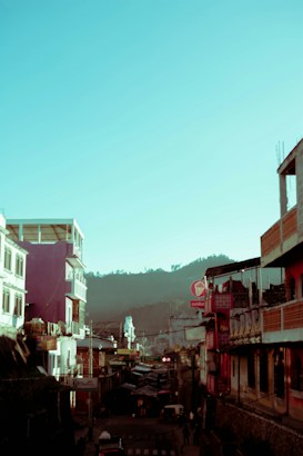 A vibrant urban street scene with colorful buildings lining both sides. The architecture shows a mix of modern and traditional styles. In the background, hazy mountains are visible under a bright blue sky, adding contrast to the bustling foreground. People and vehicles are barely discernible in the street below.