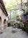 A rustic patio with stone flooring and arched stone walls. It is surrounded by lush greenery, including trees with thick branches and bushes. There is outdoor furniture, including a round table with chairs. String lights are wrapped around one of the trees, adding a cozy ambiance.