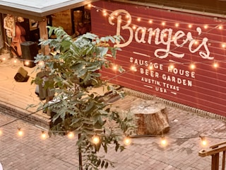 A cozy outdoor area in a beer garden with a wooden deck, stage area, and a prominently displayed sign for a sausage house called Banger's in Austin, Texas. Strings of warm lights are hanging above, creating a welcoming atmosphere. A musician is partly visible playing an upright bass.