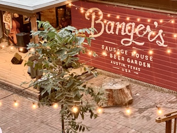 A cozy outdoor area in a beer garden with a wooden deck, stage area, and a prominently displayed sign for a sausage house called Banger's in Austin, Texas. Strings of warm lights are hanging above, creating a welcoming atmosphere. A musician is partly visible playing an upright bass.