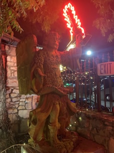 A stone statue of an angel with elaborate carvings is illuminated at night. The angel is depicted holding a neon red flaming sword that casts a glow on its intricate armor. The setting includes a stone wall, a metal fence, and an exit sign. Trees partially shade the scene, and the environment suggests an outdoor area.