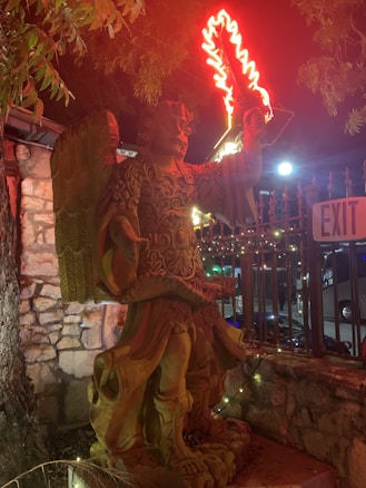 A stone statue of an angel with elaborate carvings is illuminated at night. The angel is depicted holding a neon red flaming sword that casts a glow on its intricate armor. The setting includes a stone wall, a metal fence, and an exit sign. Trees partially shade the scene, and the environment suggests an outdoor area.