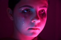 A close-up of a child's face illuminated by a pink and blue light. Glitter tears are streaked below the eyes, creating a reflective and emotional appearance. The low lighting adds dramatic shadows and highlights the glitter on the face.