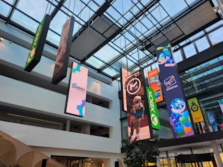 An indoor atrium with a glass ceiling showcasing multiple vertical banners hanging from the roof. The banners display various colorful graphics, including video game and technology logos such as Xbox and Godot Foundation, and animated characters. The building interior is modern with white walls and multiple floors visible.