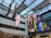 An indoor atrium with a glass ceiling showcasing multiple vertical banners hanging from the roof. The banners display various colorful graphics, including video game and technology logos such as Xbox and Godot Foundation, and animated characters. The building interior is modern with white walls and multiple floors visible.