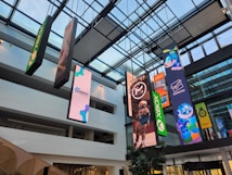 An indoor atrium with a glass ceiling showcasing multiple vertical banners hanging from the roof. The banners display various colorful graphics, including video game and technology logos such as Xbox and Godot Foundation, and animated characters. The building interior is modern with white walls and multiple floors visible.