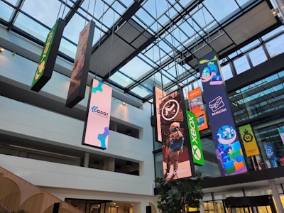 An indoor atrium with a glass ceiling showcasing multiple vertical banners hanging from the roof. The banners display various colorful graphics, including video game and technology logos such as Xbox and Godot Foundation, and animated characters. The building interior is modern with white walls and multiple floors visible.