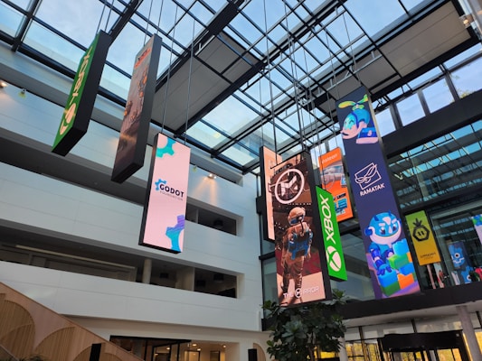 An indoor atrium with a glass ceiling showcasing multiple vertical banners hanging from the roof. The banners display various colorful graphics, including video game and technology logos such as Xbox and Godot Foundation, and animated characters. The building interior is modern with white walls and multiple floors visible.