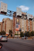 A busy urban intersection with several traffic signs mounted on an overhead structure. Behind the signs, there is an apartment building featuring a large billboard with a photo of a person and an American flag beneath it. The street is populated with cars and pedestrians, with trees lining the sidewalk.