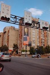 A busy urban intersection with several traffic signs mounted on an overhead structure. Behind the signs, there is an apartment building featuring a large billboard with a photo of a person and an American flag beneath it. The street is populated with cars and pedestrians, with trees lining the sidewalk.